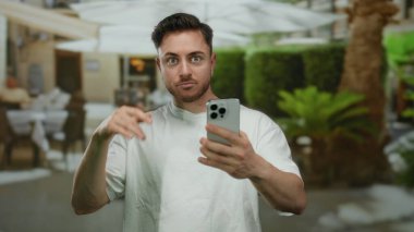 Şehirdeki açık hava restoranının terasında akıllı telefon kullanan sakallı genç adam şehir ortamında gizli bir sürpriz ve iletişim anı yakalıyor..