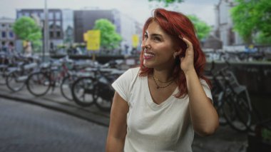 Kızıl saçlı bir kadın kulağına dokunur ve gülümser, beyaz bir tişört giyer ve bisiklet dolu bir caddede durur; kaygısız bir sevinç.