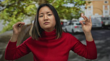Kadın, şehir caddesinde açık seçik jestler yapıyor. Arka planda bulanık arabalar ve ağaçlar var. Kırmızı süveter giyiyor, duygularını dışarıya yansıtıyor..