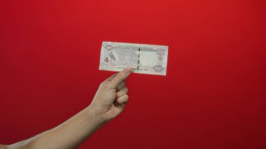 Kafkasyalı bir adam Birleşik Arap Emirlikleri 'ne bağlı Dirham Dirham banknotunu canlı kırmızı bir arka plana karşı tutuyor, izole ve odaklanmış bir şekilde para gösteriyor..