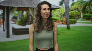 Kadın, bir binanın avlusunda dikilirken saçına dokunuyor. Palmiye ağaçlı yeşil çimlere bakıyor. Huzur..