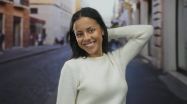 Beyaz kazaklı bir kadın, şehir caddesinde gülümserken güneşli bir gün manzarasını yakalar neşeli, canlı bir şehir anını gözler önüne serer.