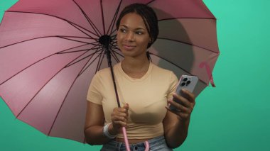 Stüdyoda pembe şemsiye ve akıllı telefon tutan kadın, bir deniz mavisi zeminine karşı ekranı okurken gülümsüyor;.