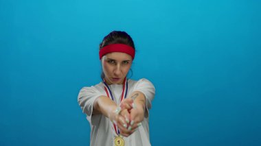 Madalyalı genç kadın, mavi arka planda kırmızı saç bandı takan, kendine güveni ve eğlencesi olan şakacı bir şekilde parmak tabancalarıyla poz veriyor..