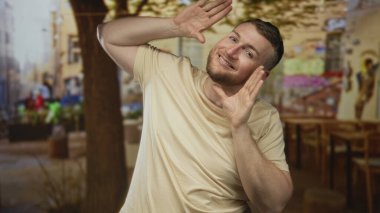 Sıcak güneş ışığı ve ağaç gölgesi altında bir sokak kafesi terasında elleri çerçeveli gülümseyen bir adam; neşeli bir an.