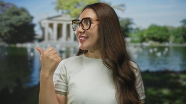 Güneş gözlüğü takan kadın, gün ışığında yeşil bir parkta sağ eliyle baş parmağını kaldırıyor..