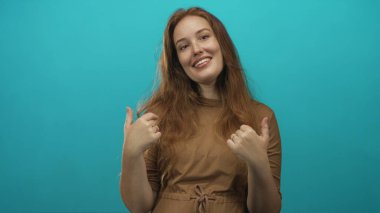 Genç gülümseyen bir kadın parlak turkuaz stüdyo sahnesinde açık avuçlar jesti sunuyor; sıcak dostça karşılama.