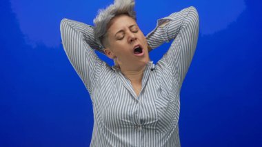 Çizgili gömlekli kadın esniyor ve kollarını başının arkasında canlı mavi stüdyo duvarına yaslıyor..