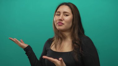 Woman holds open palms and tilts her face with a puzzled expression in studio against a teal backdrop; confusion.