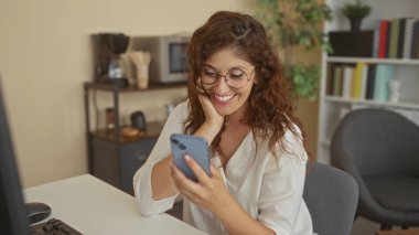 Kadın bina masasında gülümserken akıllı telefonu dinliyor, parmakları ekranda ve telefonu yüze yakın tutarken; mutluluk ve bağlantı.