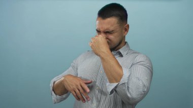 Çizgili gömlekli adam mavi stüdyo duvarına karşı burnu sıkıyor, gözleri kapalı, diğer eli havada;.