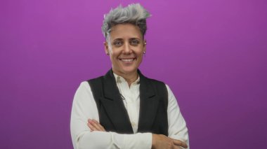 Kısa gri saçlı, gülümseyen ve kollarını kavuşturan bir kadın stüdyoda canlı mor arkaplanlı, kendine güvenen..