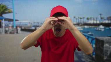 Kırmızı şapkalı genç adam dürbünle bakıyor ve ellerini sokakta gezdiriyor..