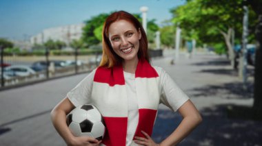 Şehir caddesinde elinde futbol topu tutan kırmızı beyaz eşarp takan neşeli şehir ortamında gülümseyen bir kadın..