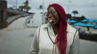 Sahilde kırmızı örgülü Afro-Amerikalı bir kadın, neşeli bir dille, kumsalda güneşli bir günün tadını çıkarıyor parlak şemsiyeler ve sakin bir arka planla.