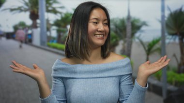 Deniz kenarındaki bir güvertede neşeyle gülümseyen bir kadın, neşeli yüzünü palmiye ağaçları ve mavi gökyüzü ile sergiliyor, dingin çevreyi büyütüyor..