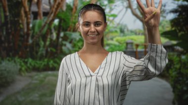 Yeşil yaprakların yanında sarı tırnaklı, gülümseyen ve sağ elini sallayan kadın, beyaz çizgili bluz giyiyor ve yükseltilmiş palmiye gösteriyor; sevinç selamlaması.