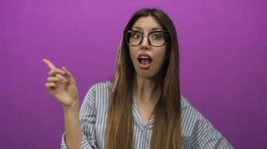 Gözlük takan ve çizgili gömlek giyen genç bir kadın stüdyoda mor duvara parmak işareti yapıyor..