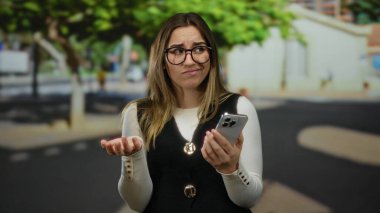 Gözlüklü bir kadın güneşli bir sokakta akıllı telefon kullanıyor. Arka planda ağaçlar ve binalar olan sıradan bir açık hava ortamında düşünceli görünüyor..