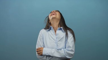 Kolları çapraz gülen, stüdyoda dişlerini mavi duvarla gösteren açık mavi gömlek giyen neşeli, spontane bir kadın..