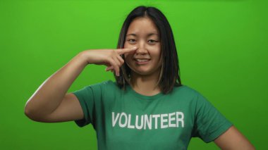 Yeşil arka planda gönüllü gömleği olan bir kadın burnuna işaret ediyor, neşeli bir ifade sergiliyor ve tek başına ayakta duruyor..
