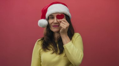 Sarı bluz giyen ve Noel Baba şapkalı bir kadın kırmızı arka planda kırmızı bir kalp taşıyor. Noel sezonunda neşeli tezahürat ve şefkat gösteriyor..