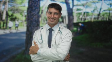 Steteskoplu beyaz önlüklü adam ağaçlarla dolu yeşil parkta gülümserken kollarını kavuşturuyor..