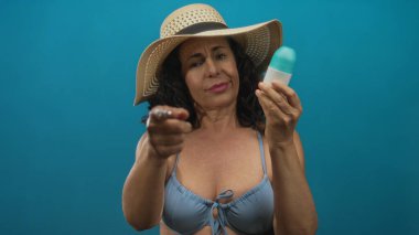 Şapka ve bikini giyen bir kadın, izole edilmiş mavi arka planda losyon şişesini inceliyor, rahat bir yaz havası sürdürürken meraklı bir ifade yayıyor..