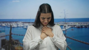 Elleri göğsünde, arka planda teknelerle limanda duran, beyaz gömlek giyen, açık denizde duygusal ve yansıtıcı bir an yaşayan bir kadın..