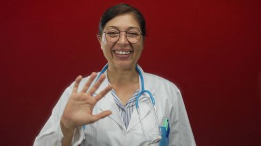 Orta yaşlı İspanyol doktor beyaz önlük giyiyor kırmızı stüdyoda steteskopla el sallıyor; empati.