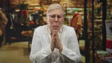 Beyaz gömlek ve gözlüklü bir kadın, bir giyim mağazası içinde el ele yalvarma jest bastırılmış; savunma umutsuzluk.