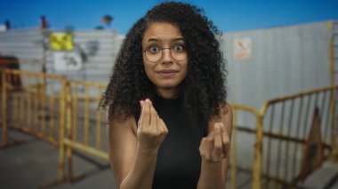 Genç bir kadın ellerini sarı bir bariyerin yanında birbirine bastırır ve bina girişini açık havada yapar; çaresizlik.