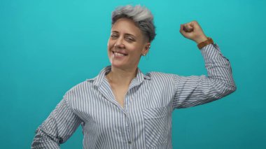 Çizgili gömlekli kadın, mavi stüdyoda pazı kasını göstermek için kolunu esnetir; kendine güvenin dayanıklılığını güçlendirir.