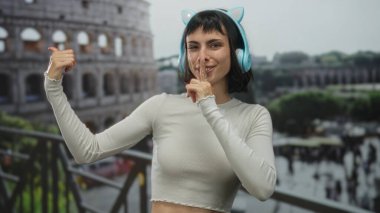 Kolezyum binasında sessiz kalmak için kedi kulaklığı ve parmak dudağa konuşan Latin kökenli genç bir kadın..