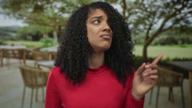 Afrika kökenli Amerikalı bir kadın, güneş ışığı altında restoran terasına şüpheyle bakan kırmızı gömleği işaret ediyor..
