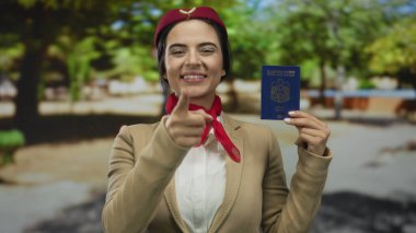 Genç İspanyol uçuş görevlisi üniformalı gülümseyip işaret ederken Birleşik Arap Emirlikleri pasaportunu açık mavi gökyüzünün altında tutuyor..