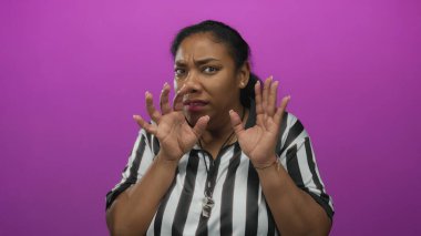 Çizgili gömlek giyen kadın hakem, mor stüdyo duvarına karşı ıslık çalmanın yanında el kaldırıyor, siniyor ve kaşlarını çatıyor..