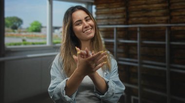 Geniş bir pencere ve ahşap merdiven duvarının yakınındaki kapalı bir stüdyoda el çırpan kadın kameraya doğru gülümsüyor..