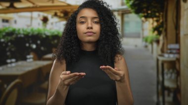 Bir restoranın terasında duran kıvırcık saçlı, elleriyle hareket eden, siyah giyen, gündüzleri dışarıda gezen, duygularını ve ifadesini sergileyen bir kadın..