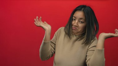 Kafası karışmış bir kadın omuzlarını kırmızı bir arka plana doğru silkiyor, belirsizlik ve düşüncelere dalmış bir şekilde tecrit edilmiş sahnede yaşıyor..