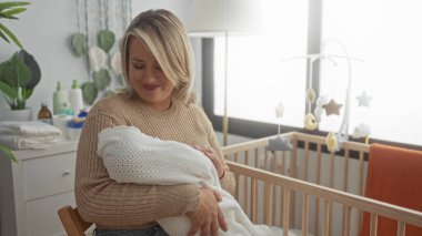 Stüdyoda ahşap beşiğin yanında beyaz battaniyeye sarılı uyuyan bebeği nazikçe kucaklar; anne sevgisi dinginliği.