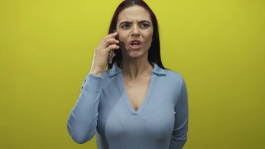 Telefondaki kadın canlı sarı bir duvara karşı yoğun bir ifadeyle konuşuyor, mavi bir gömlek giyiyor, hayal kırıklığı ve iletişim anını yakalıyor..