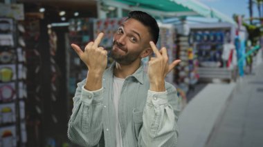 Deniz kenarındaki gezinti güvertesinde gülümseyen genç adam, canlı bir şekilde el hareketleri yapıyor. Parlak dükkan dekorları ve güneşli atmosferle birlikte..