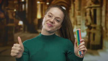 Yeşil kazaklı gülümseyen kadın bir binanın içinde başparmak hareketi yaparken renkli kalemler tutuyor; yaratıcılık.