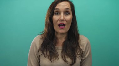 Canlı yeşil arka planda gülümseyen bir kadın uzun siyah saçlı ve bej renkli kazaklı neşeli ve şaşırtıcı ifadeler sergiliyor..