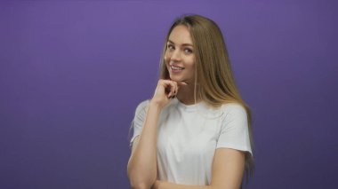 Mor arka planda beyaz gömlekli bir kadın düşünceli bir gülümsemeyle düşünceli, izole ve sakin bir ifadeyle elleri rahat bir duruşla çenenin altında dinleniyor..