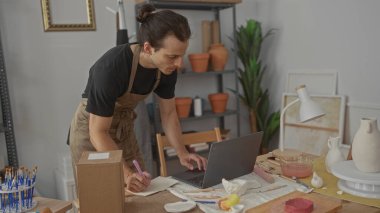 Önlüklü bir adam dizüstü bilgisayarının yanında kalemle yazı masasına yaslanıyor ve atölyedeki çalışma masasında kilden vazolar bulunuyor..