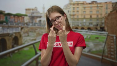 Kırmızı gömlekli gönüllü kadın, Roma harabelerine tepeden bakan antik binada şans diliyor; iyimserlik dayanışması.
