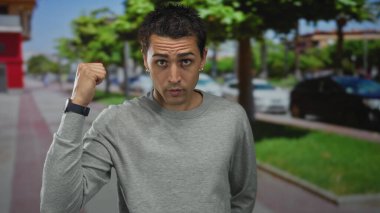 Gri kazaklı genç İspanyol adam şehir caddesinde ağaçlı ve park edilmiş arabalı kamerayı işaret ediyor..