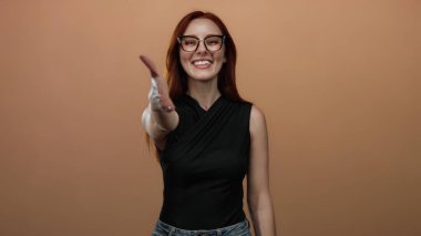 Dost canlısı bir şekilde gülümseyen bir kadın basit bir kahverengi arka plana karşı gözlüklü ve kendinden emin bir ifadeyle günlük siyah kıyafetli..